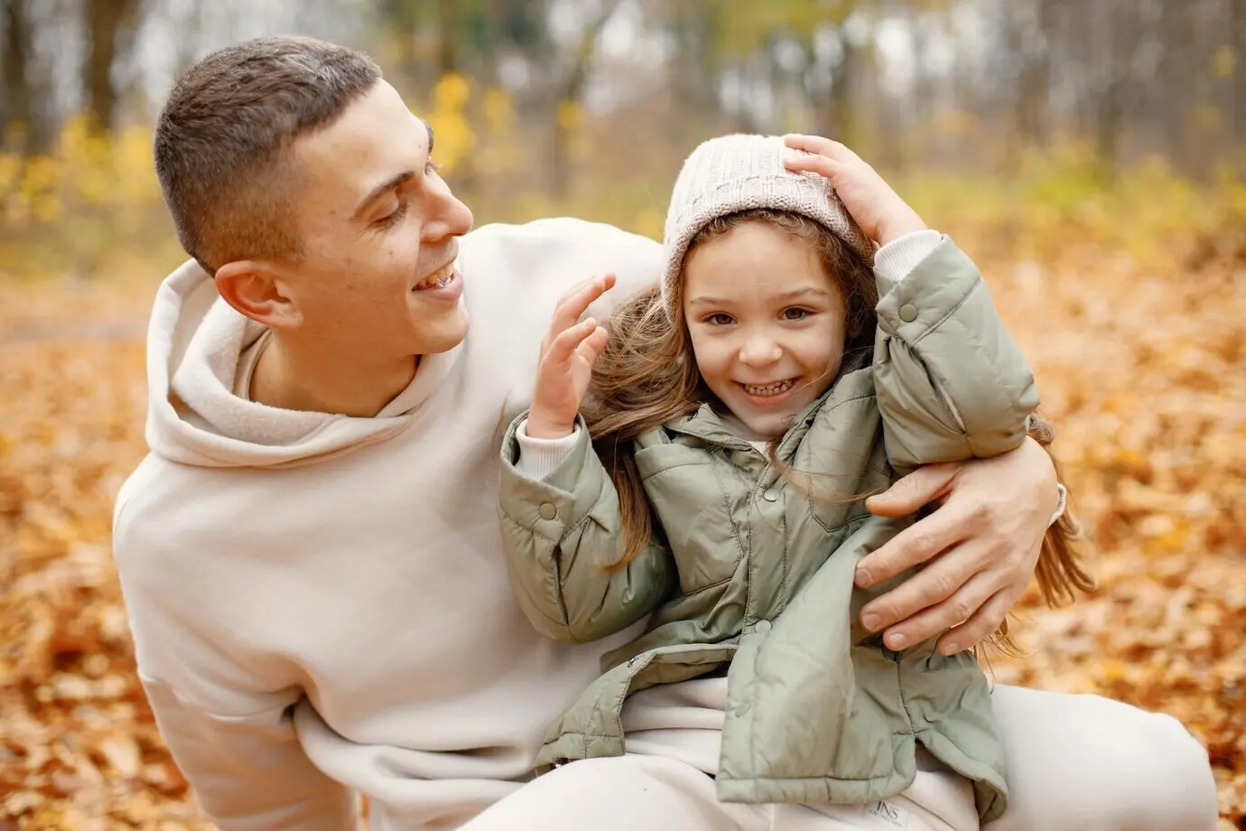 Ein Mann und sein kleines Mädchen spielen lachend im herbstlichen Wald. Der brünette Mann und das kleine Mädchen umarmen sich. Die Familie trägt beige sportliche Kleidung.